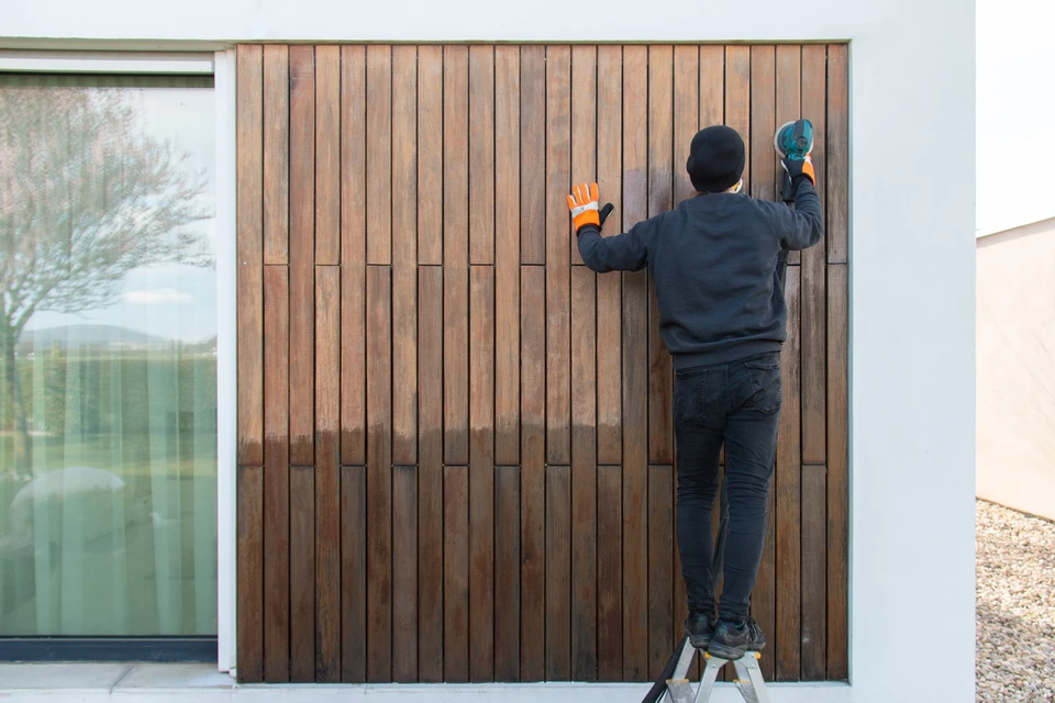 Un ouvrier en vêtements sombres et gants orange installe un bardage bois vertical sur la façade extérieure d'une maison moderne