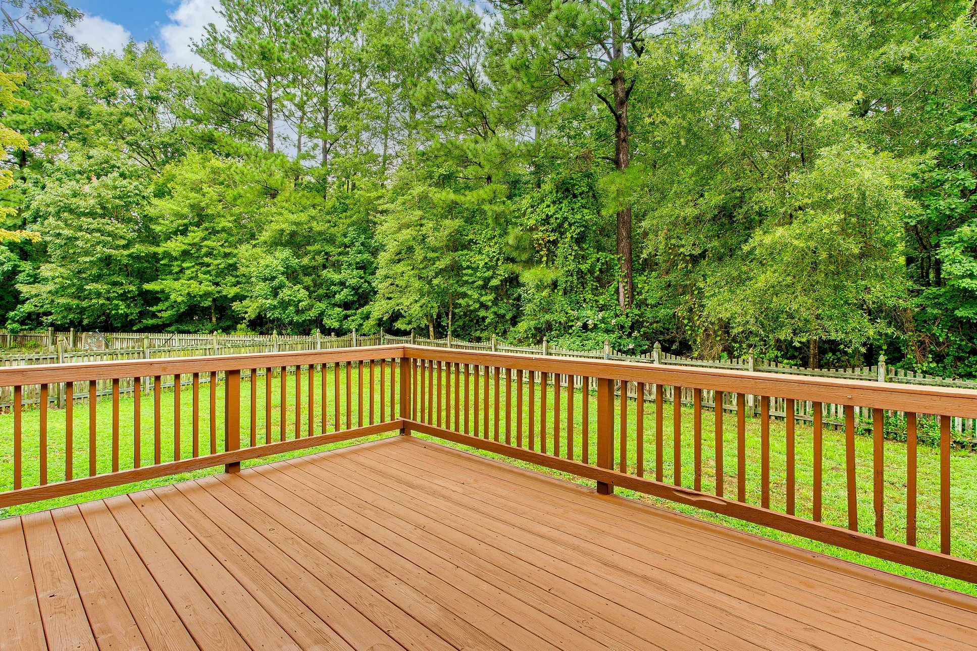 Terrasse extérieure en bois avec garde-corps à barreaux verticaux donnant sur jardin verdoyant et forêt dense d'arbres feuillus