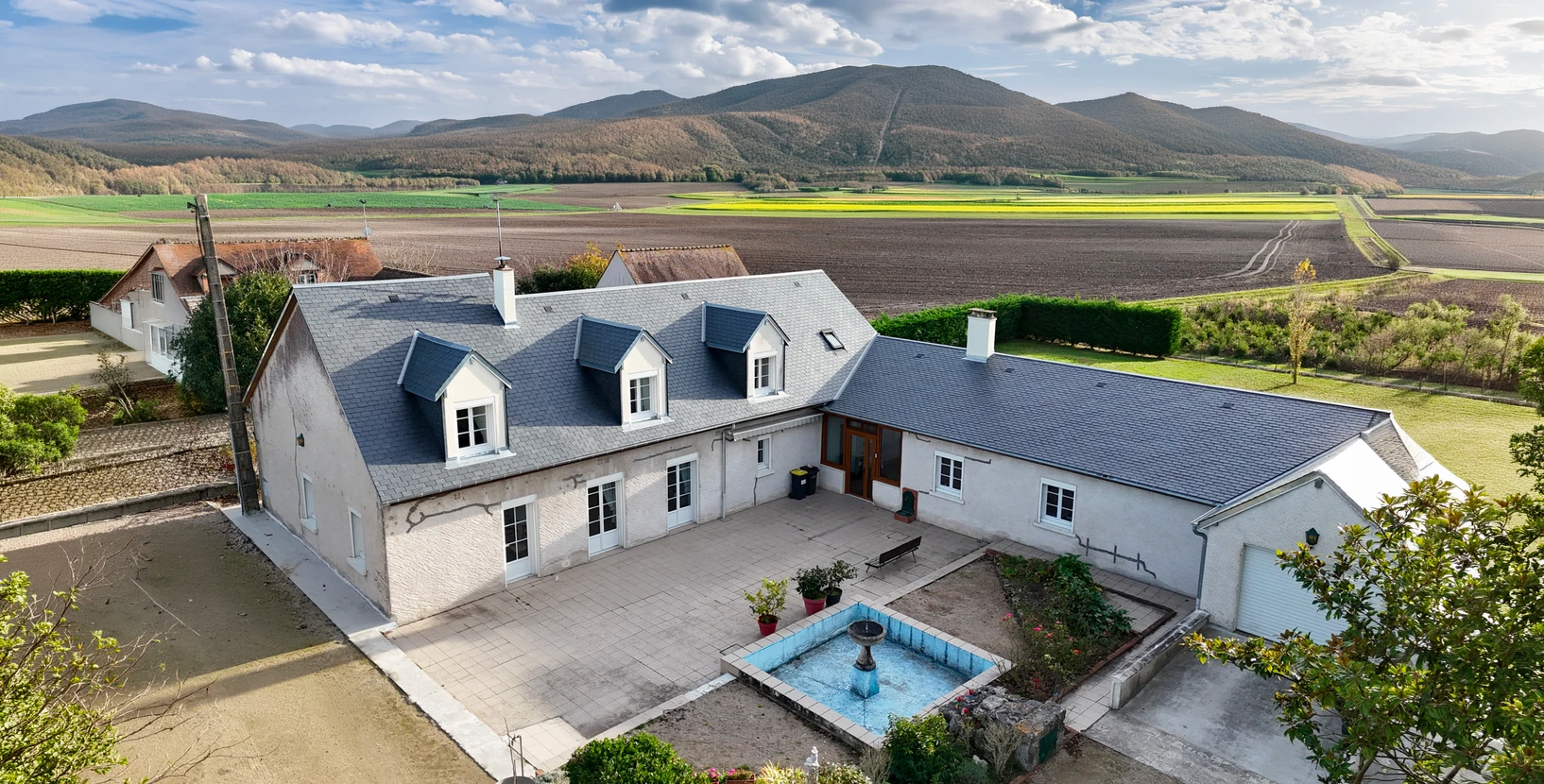 Vue a&eacute;rienne par drone d'une propri&eacute;t&eacute; de caract&egrave;re avec cour int&eacute;rieure pav&eacute;e, fontaine centrale carr&eacute;e, toiture en ardoise bleue avec lucarnes &agrave; frontons, murs blancs