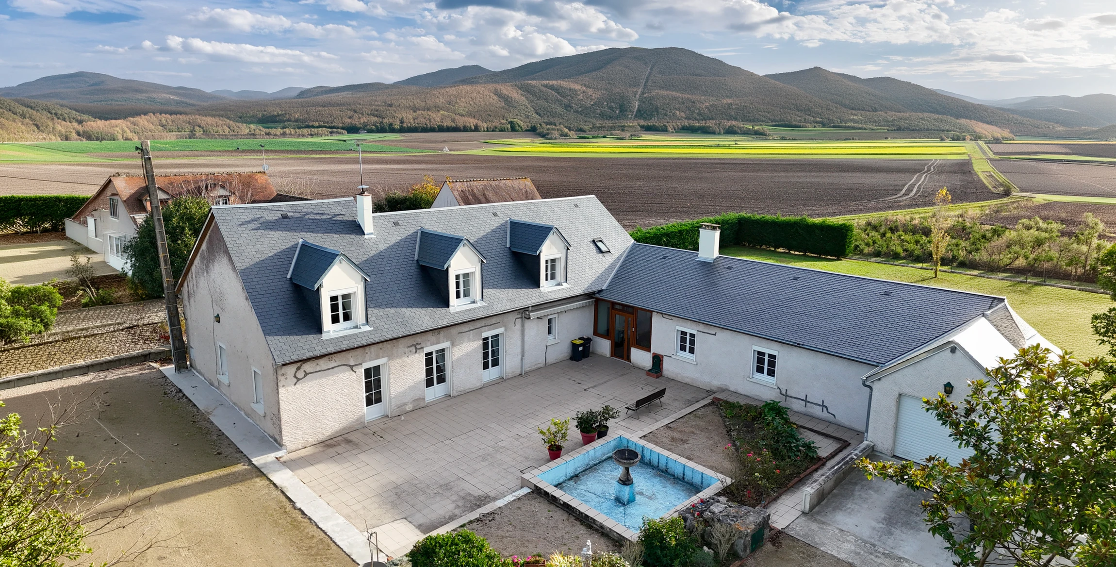 Vue aérienne par drone d'une propriété de caractère avec cour intérieure pavée, fontaine centrale carrée, toiture en ardoise bleue avec lucarnes à frontons, murs blancs